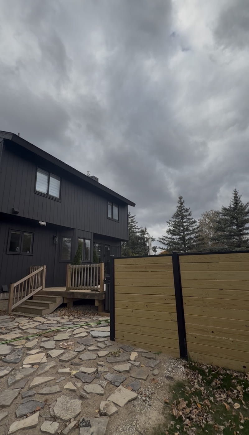 Completed dark charcoal board-and-batten siding on Royal Oak home with wood deck, horizontal fence, and flagstone patio